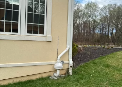 White radon mitigation system installed outside a beige house beside a window on a grassy lawn.