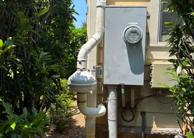Gas meter and electrical box mounted on an exterior wall, surrounded by green shrubs and a dirt path.