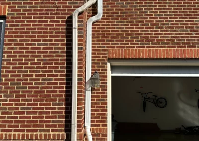 White pipes run vertically along a red brick house wall beside an open garage with bikes visible inside.