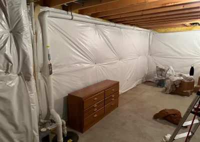 Unfinished basement with exposed beams, white plastic-covered walls, a wooden dresser, and scattered items.