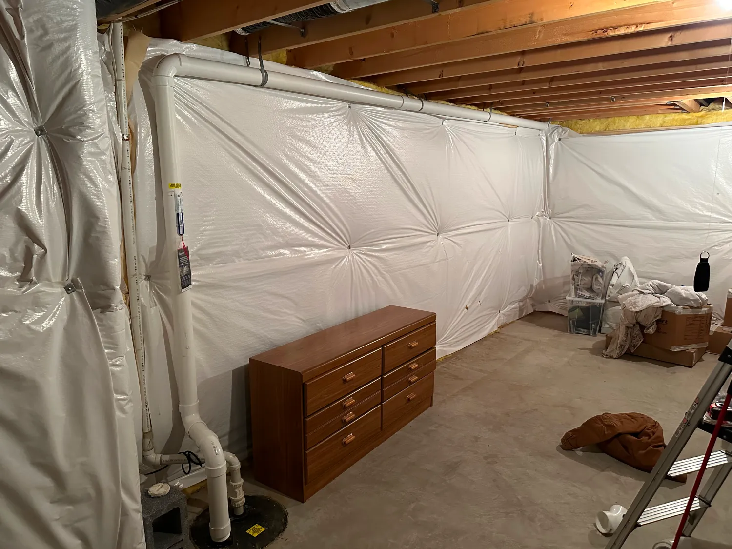 Unfinished basement with exposed beams, white plastic-covered walls, a wooden dresser, and scattered items.