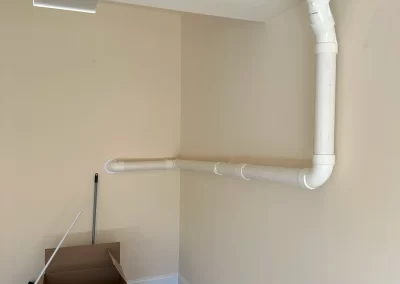 White PVC pipes run along a beige wall in a room; an open box and broom are on the bare floor below.