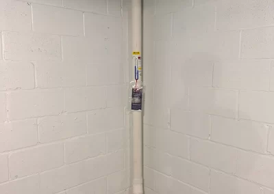 A white PVC pipe runs vertically in the corner of a basement with painted cinder block walls.