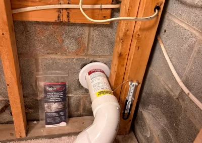 White PVC radon mitigation pipe installed in a basement corner, next to a radon system gauge and info sheet.