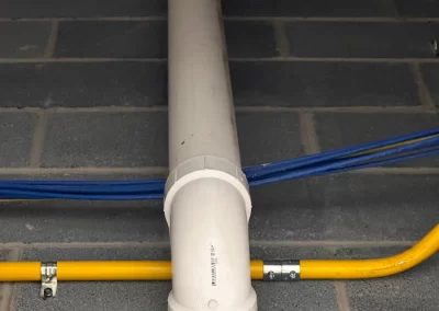 White PVC pipe with an elbow joint runs vertically along a cinder block wall, with blue and yellow cables nearby.