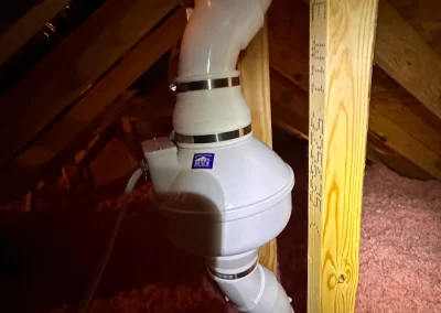 A white radon mitigation fan attached to PVC pipes in an attic with wood framing and pink insulation.