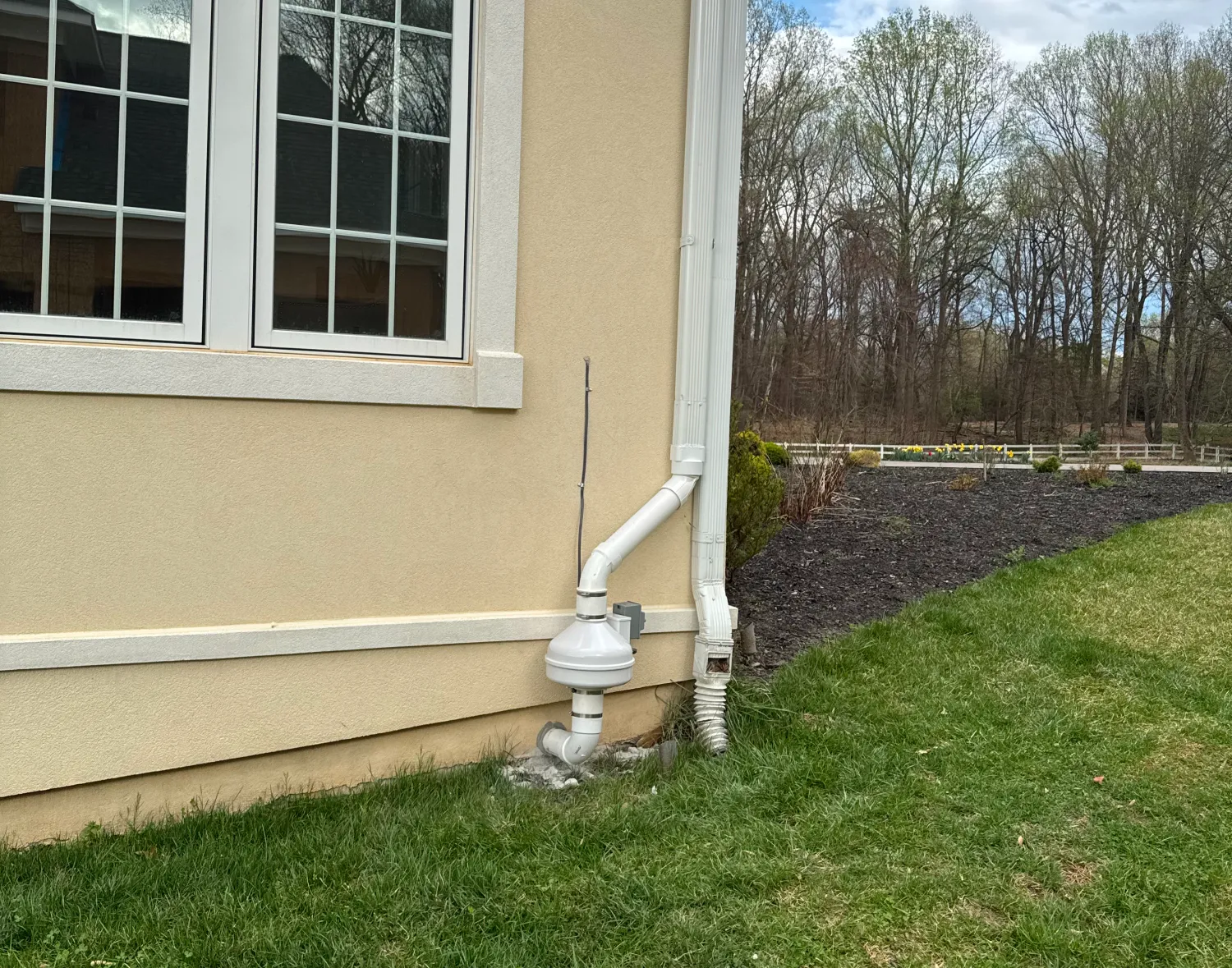 White radon mitigation system installed on the exterior wall of a beige house near a grassy yard.
