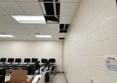 A classroom with missing ceiling panels, exposed pipes, computers, boxes, and a vacuum on the floor.