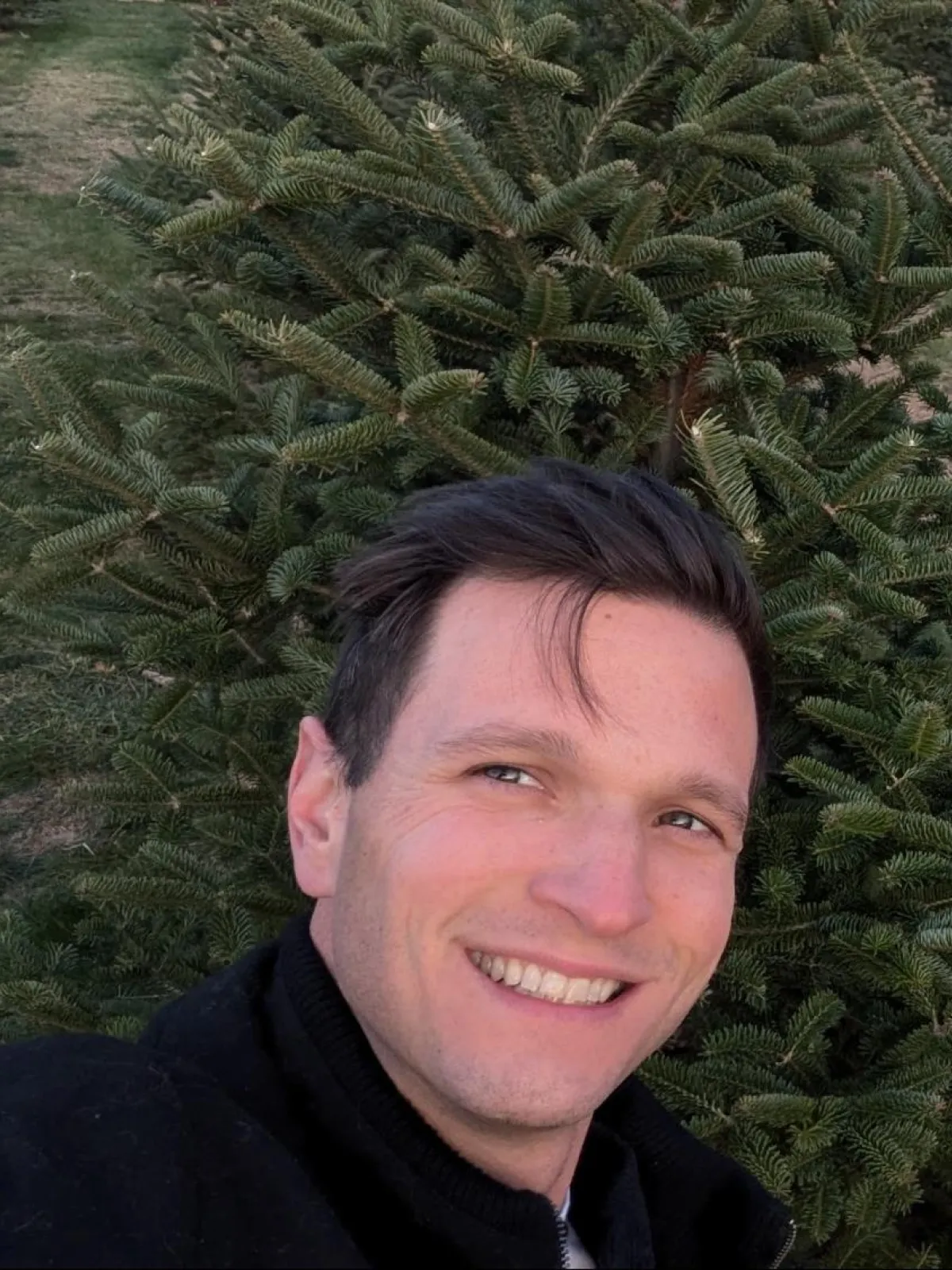 Smiling person taking a selfie outdoors in front of a green pine tree.