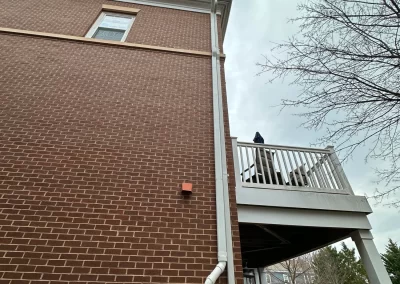 A brick building with a white downspout and a person standing on a balcony near a parked car.