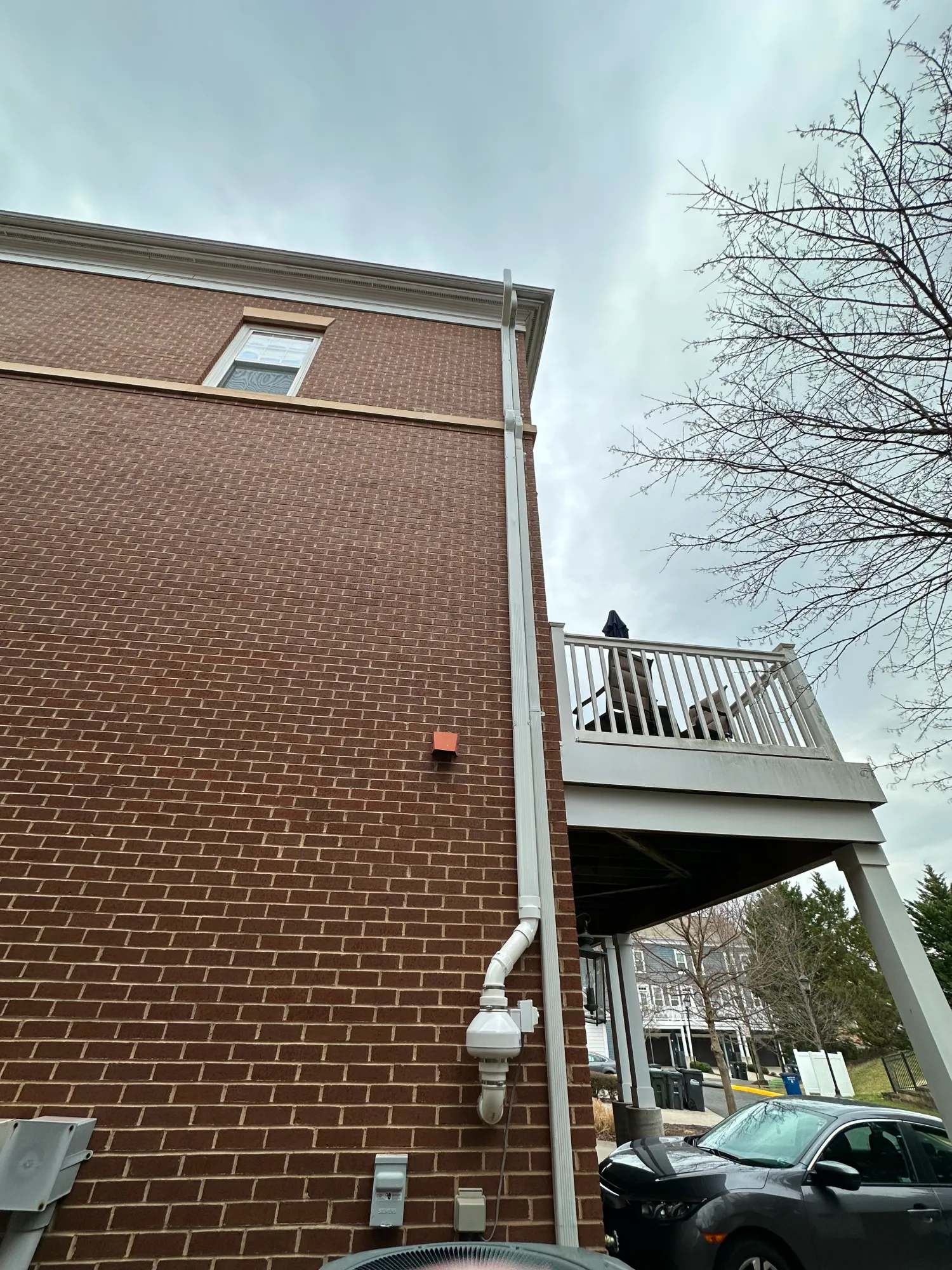 A brick building with a white downspout and a person standing on a balcony near a parked car.