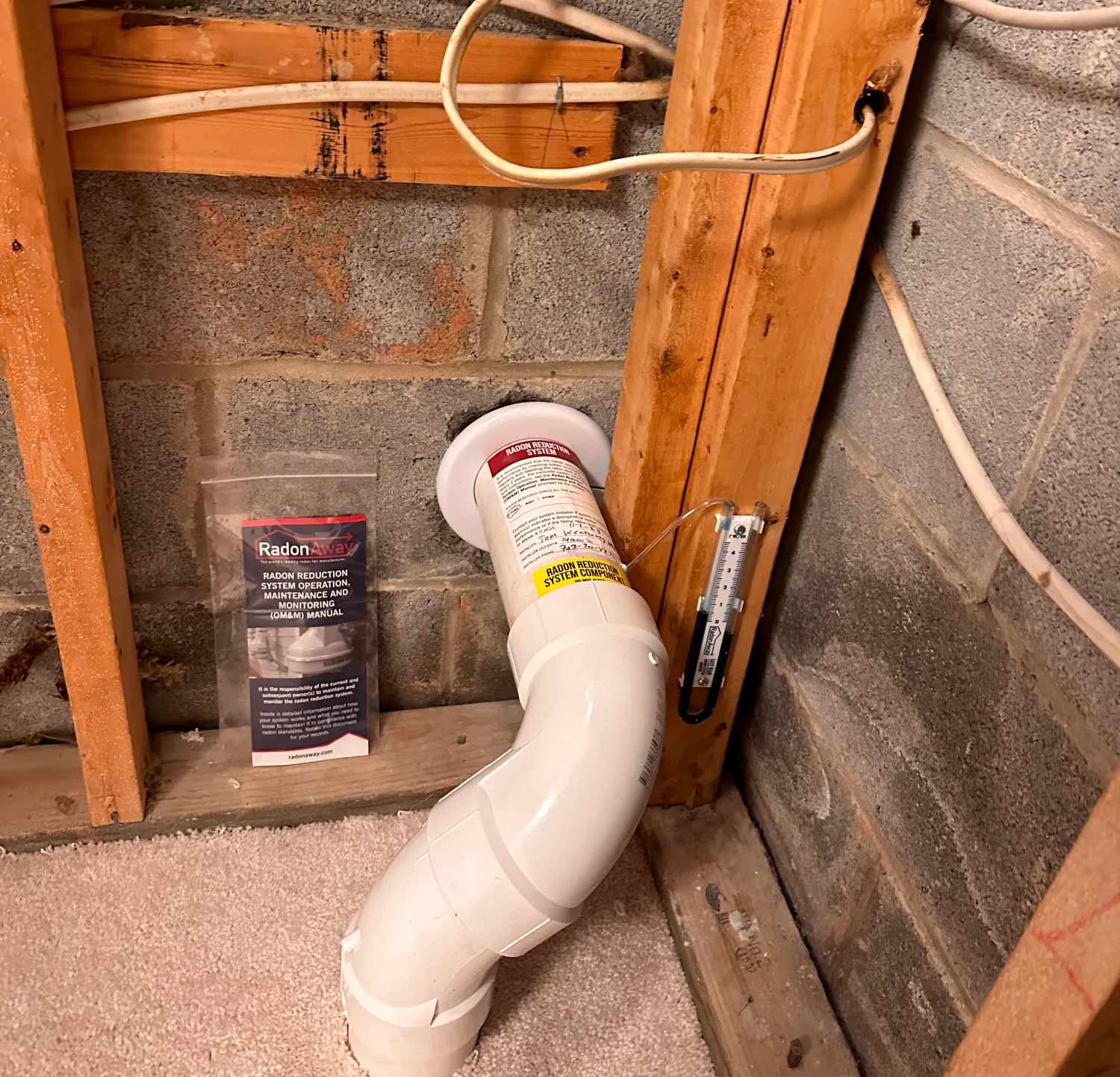 A radon mitigation system pipe with a warning label installed in a basement near a bagged radon test kit.