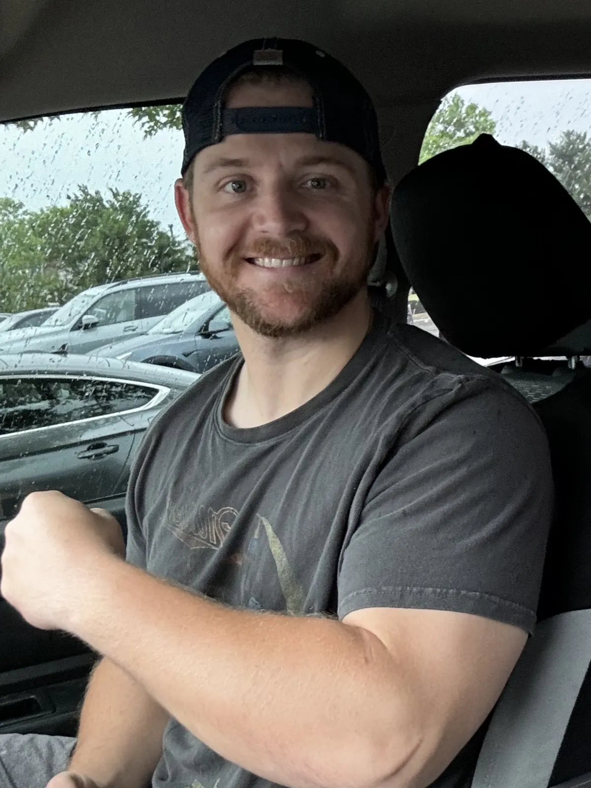 Smiling man in a dark t-shirt and cap sitting in a car on a rainy day, pointing to his left.