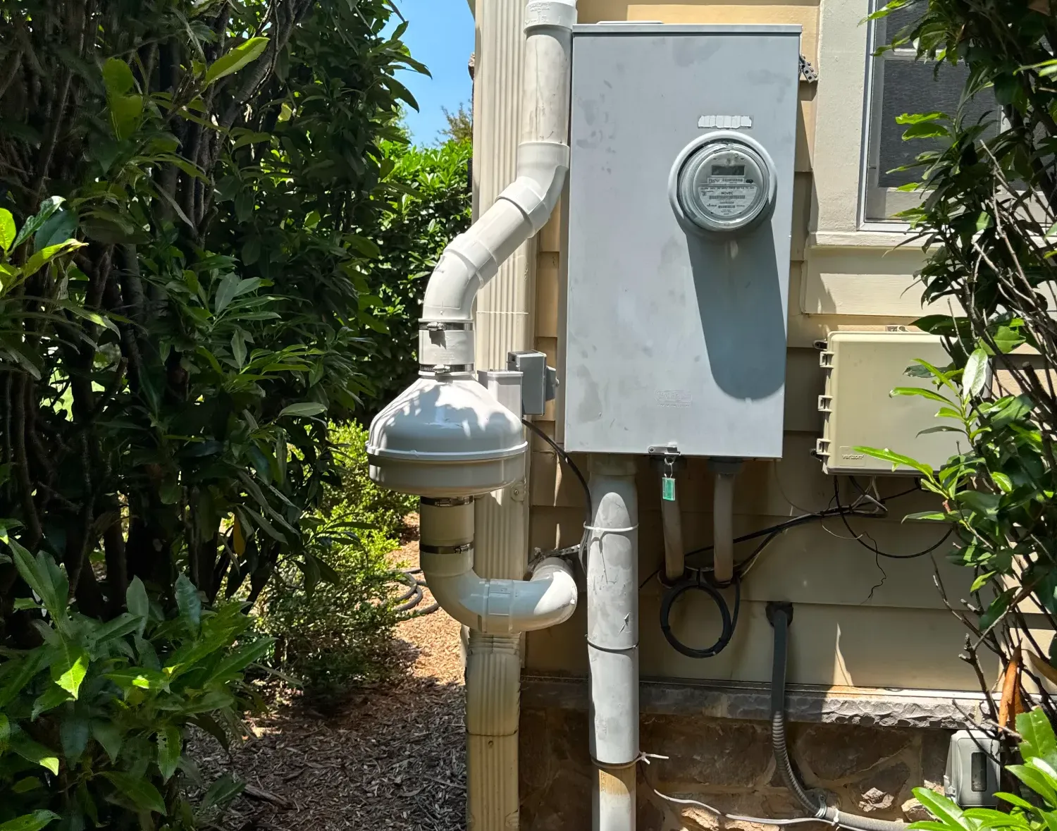 Outdoor radon mitigation system with pipes and a fan installed next to an electric meter on a house wall.