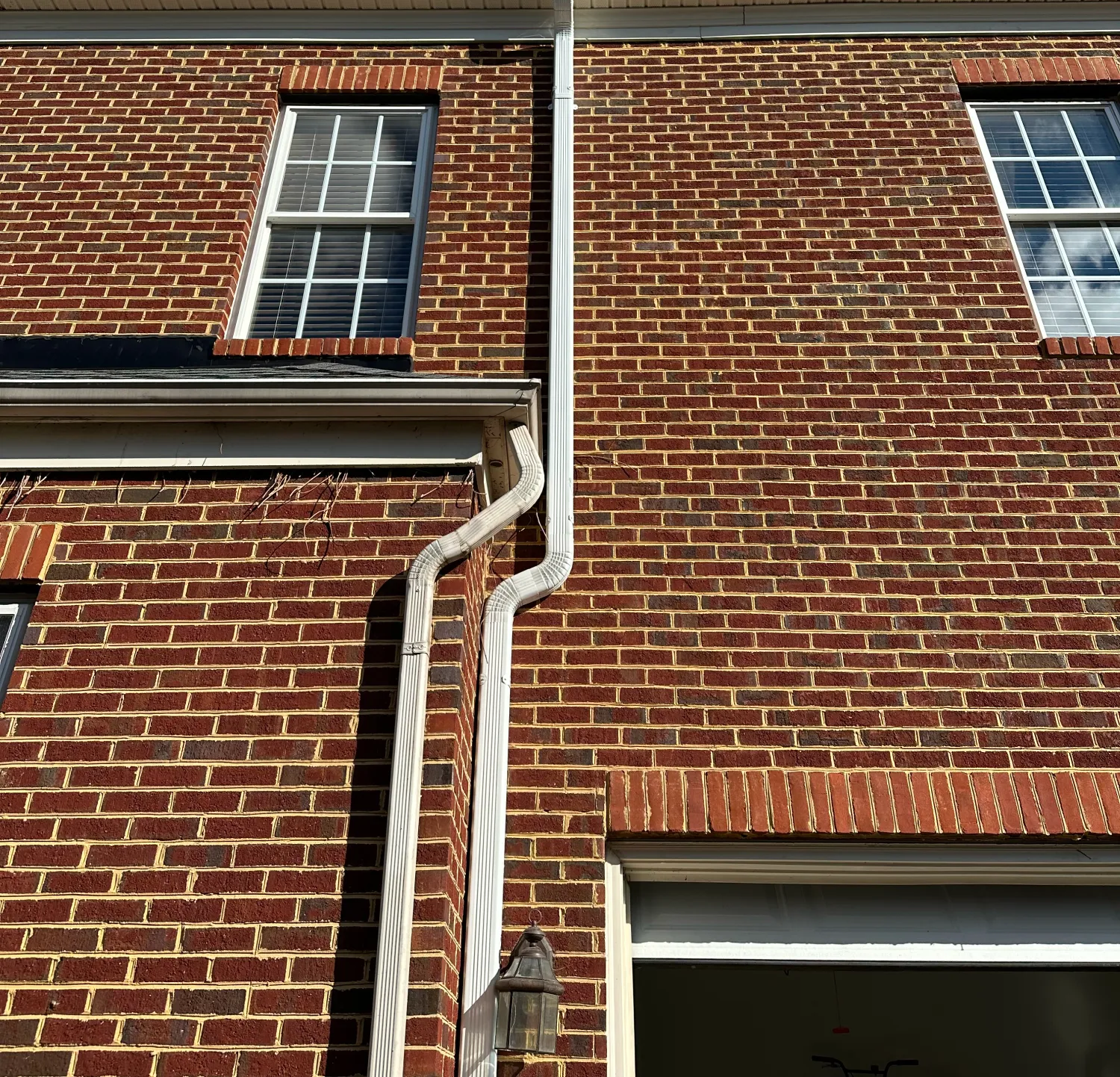 White gutter downspout with multiple bends attached to a red brick house near windows and a garage.