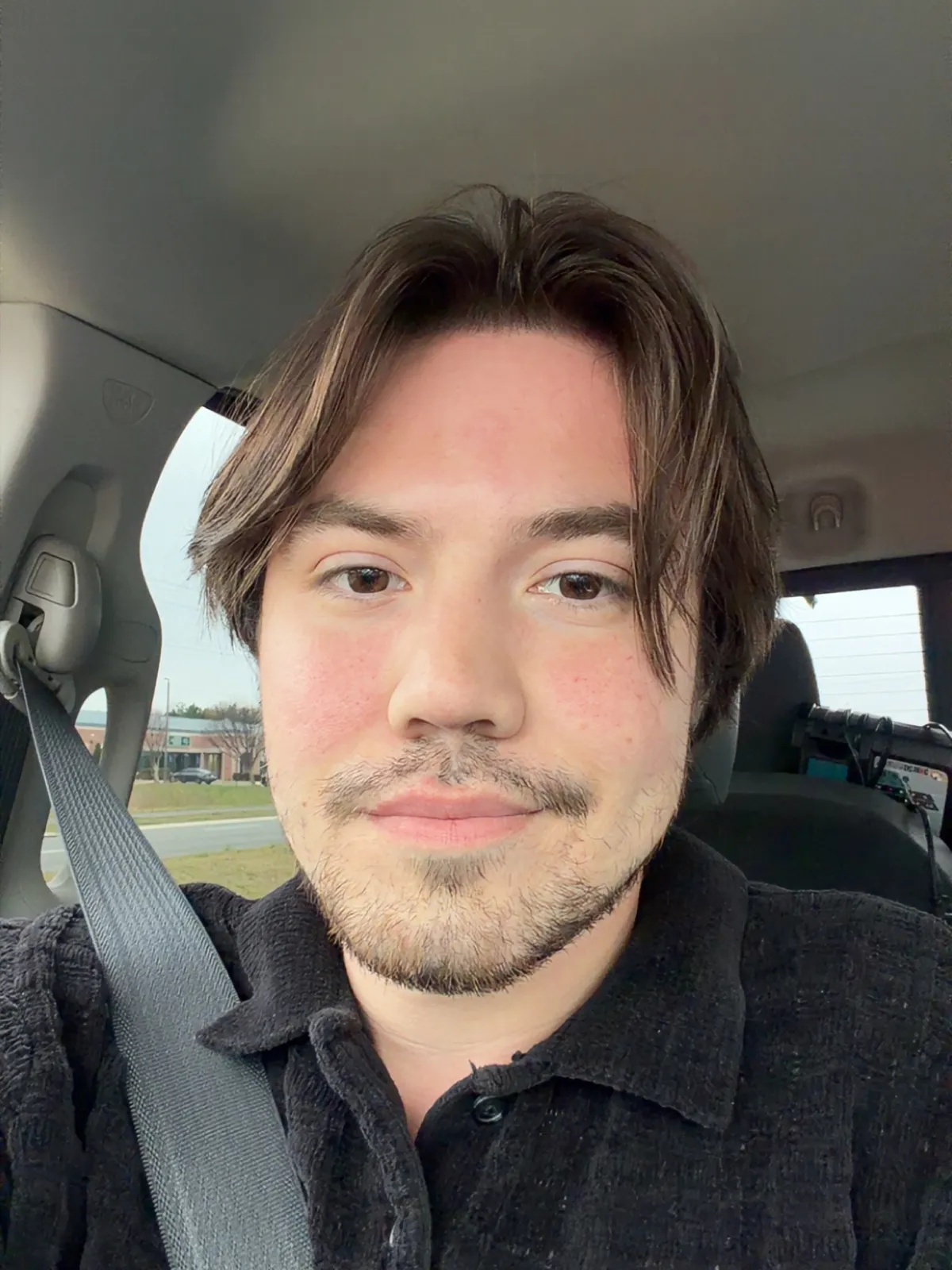 A young man with brown hair and a beard sits in a car, wearing a seatbelt and a black shirt.